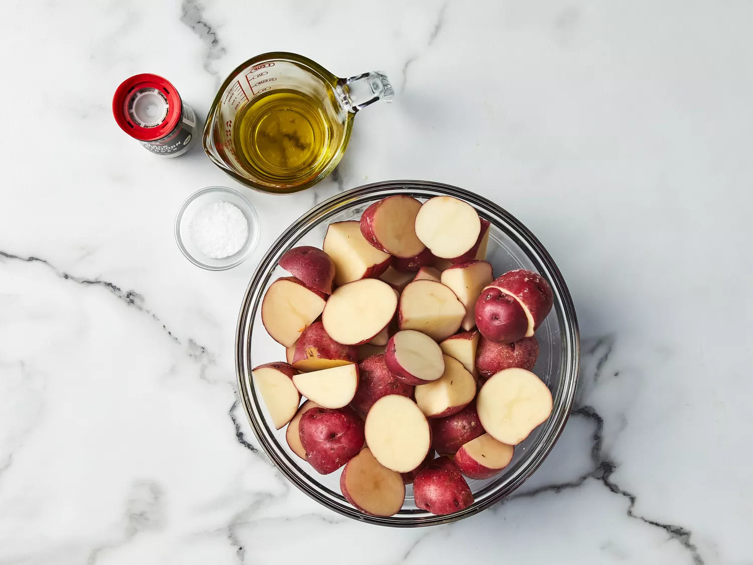 Red potatoes halved in a bowl with olive oil, salt, and pepper on the side